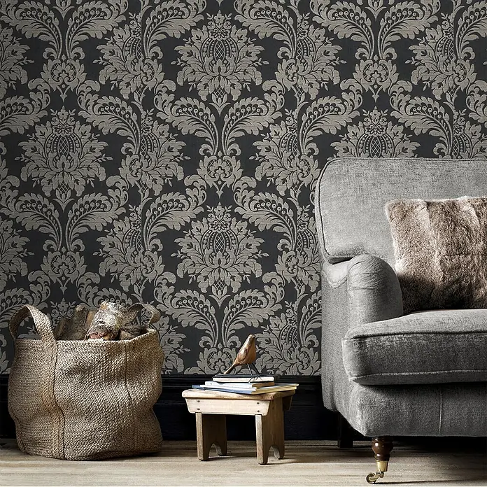 Sublime Vliestapete Damask - Schwarz - Dunkle anthrazitfarbene Tapete mit elegantem metallisch-grauem Damastmuster, verschönert ein Wohnzimmer neben einem grauen Sessel.