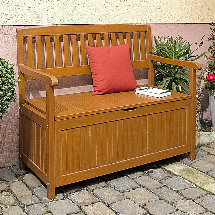 Sunfun Diana Gartenbank - L x B x H: 107 x 57 x 90 cm, Eukalyptus - Eine braune Gartenbank aus Holz mit Stauraum, einem roten Kissen und einem Buch darauf vor einer Wand.