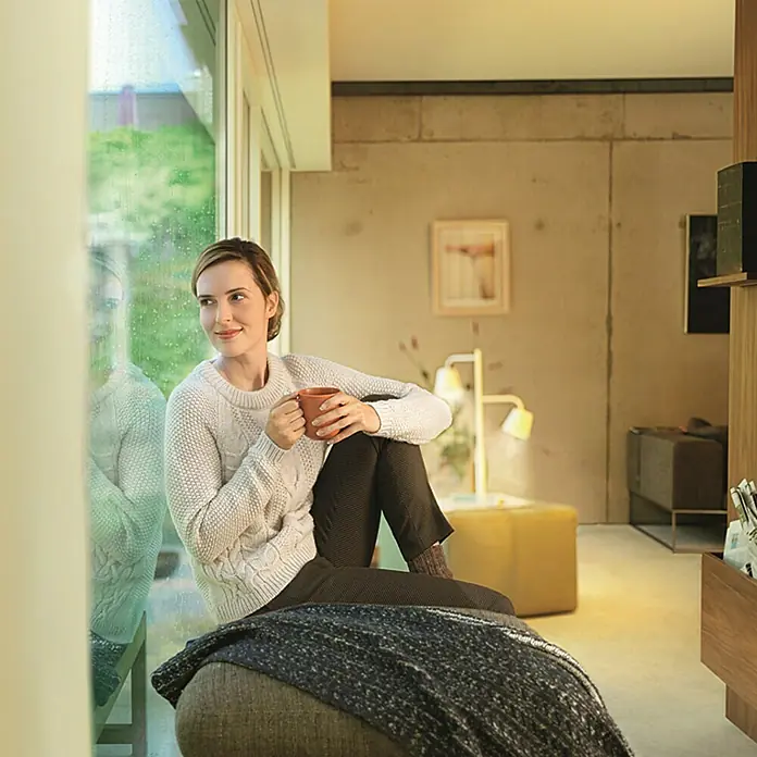 Osram Lightify Bombilla LED Classic A60 - E27, Intensidad regulable, Blanco neutro, 310 lm, 9,5 W - Una mujer sentada junto a la ventana disfruta de una taza de café. La vista destaca la acogedora y moderna sala de estar con colores cálidos.