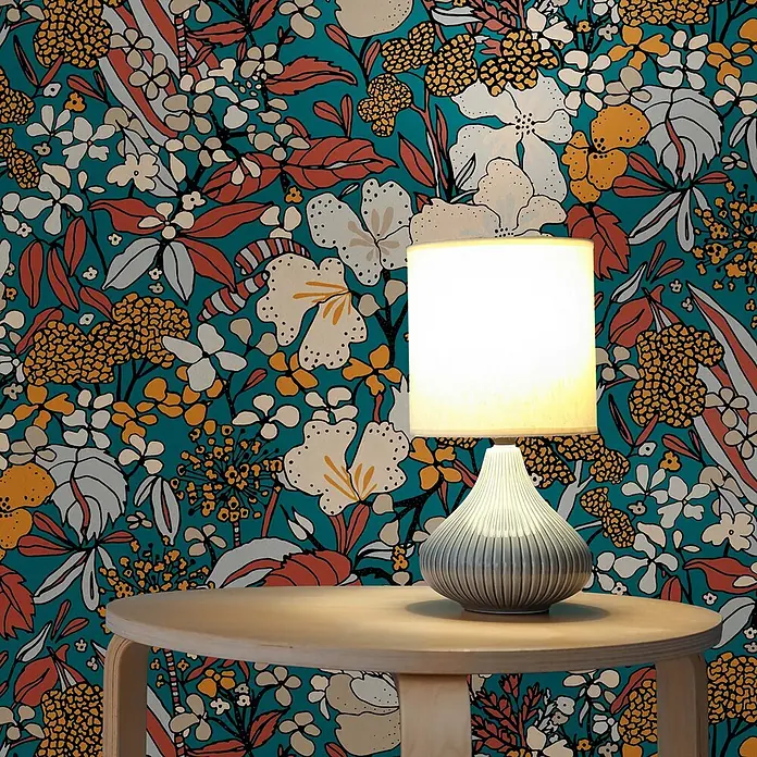 Tapete Dschungel Aleix M1-No.565 Vliestapete Bunt Gelb Natur Palmen -  - Eine leuchtende Tischlampe mit weißem Schirm und strukturiertem grauem Fuß steht auf einem hellen Holztisch vor einer floralen Tapete.
