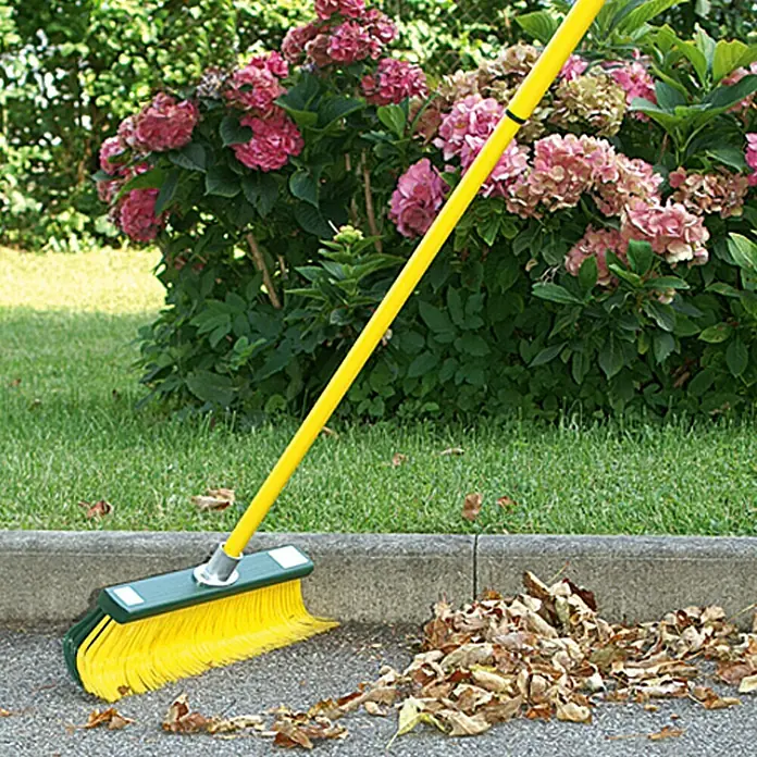 Steuber Krallenbesen - Breite: 45 cm, Einsatzbereich: Garten - Gelber Besen mit grünem Kopf beim Aufkehren von Blättern auf einer asphaltierten Fläche vor einem Garten.