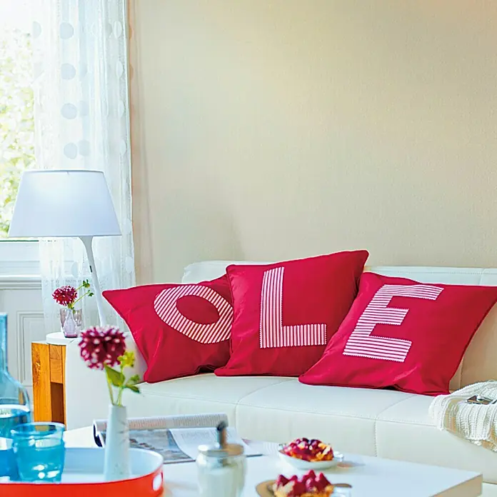 SCHÖNER WOHNEN-Farbe Wandfarbe Trendfarbe - Cashmere, Matt, 1 l, Konservierungsmittelfrei - Dekorative rote Kissen mit weißen Buchstabenaufdrucken liegen auf einem weißen Sofa, das eine einladende Atmosphäre schafft.