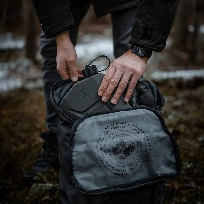 Oxid7 by BW Kraftstoffkanister Proper 2 l, Metall + Ausgießer, 2 Stk. -  - Eine Person legt eine dunkelgrüne 2L Feldflasche in einen schwarzen Rucksack.