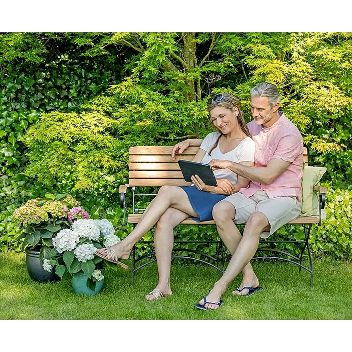 Teak Gartenbank Stahl und Teakholz 2-Sitzer Klappbank wetterfest edel -  - Holzgartenbank mit dunklem Metallgestell, von einem Paar zur Entspannung im Garten genutzt.