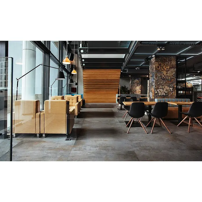Feinsteinzeug in grauer Betonoptik 120x120 - Italienische Fliesen -  - Modernes Café-Interieur mit beigen Loungesesseln, dunklen Esstischen, Holzwand und Graffiti-Säulen. Helle Fenster.