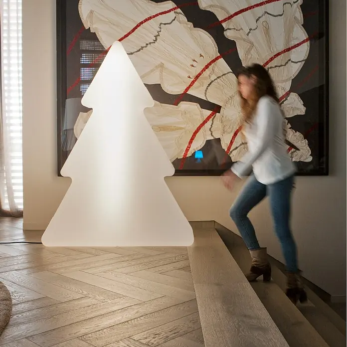 New Garden Árbol de Navidad de LEDs Pinus - Para exterior, 160 cm, 1 ud., Color de luz: Blanco neutro, Funcionamiento en red - Lámpara de pie grande, blanca y translúcida con forma de árbol de Navidad que ilumina una habitación, con una persona pasando.