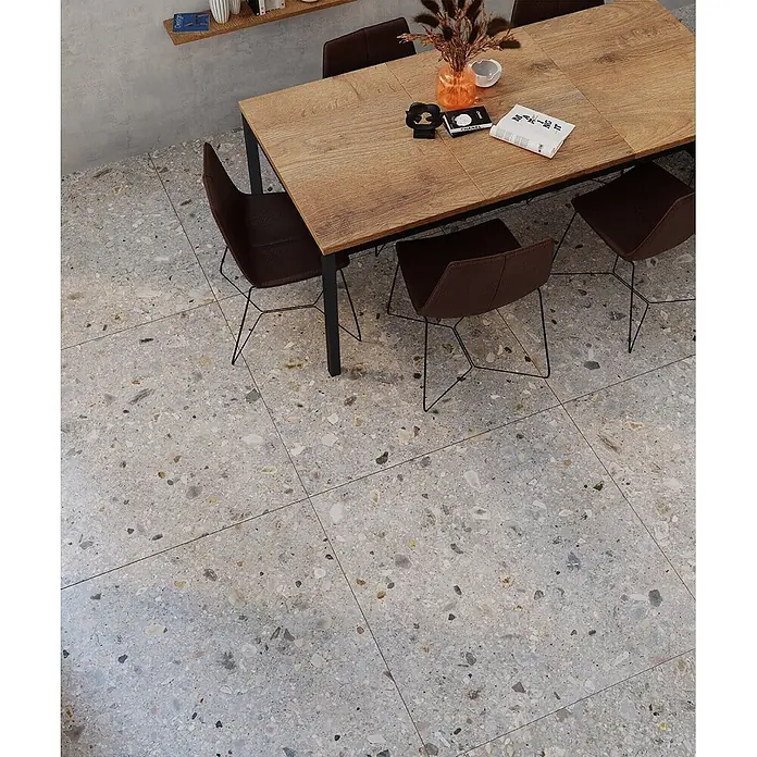 Feinsteinzeug in matter hellgrauer Steinoptik 120x120 - Ital. Fliesen -  - Große graue Terrazzo-Fliesen mit bunten Chips, verlegt als Esszimmerboden unter einem Holztisch und braunen Stühlen.