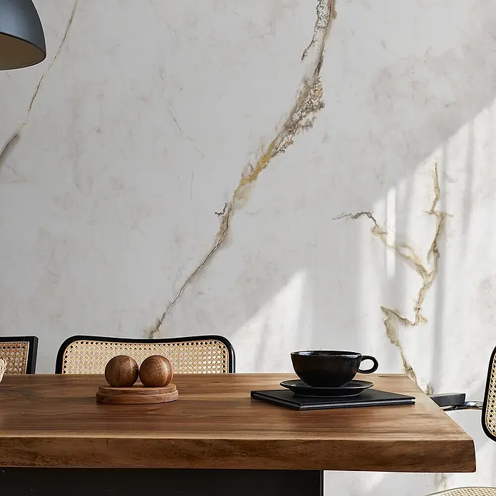 Feinsteinzeug in Gold extra matter Calacatta-Marmoroptik 60x120 -  - Ein modernes Esszimmer mit einem langen Holztisch, Stühlen mit Korbstofflehnen und einer marmorierten Wand. Eine schwarze Tasse und Holzdekorationen stehen auf dem Tisch.
