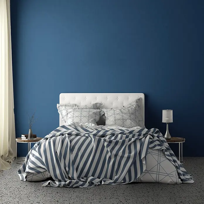 Feinsteinzeug in farbiger dunkelblauer Betonoptik 60x60 - Ital. Fliesen -  - Gemütliches Schlafzimmer mit weißem Bett, gemusterten Kissen und blau-weiß gestreifter Decke vor einer blauen Wand.