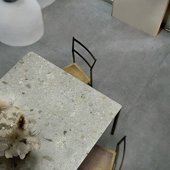 Feinsteinzeug in moderner dunkelgrauer Betonoptik 80x80 - Ital. Fliesen -  - Blick von oben auf einen rechteckigen Tisch im Terrazzo-Stil mit Stühlen, auf einem grauen Fliesenboden.