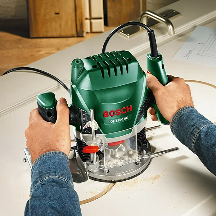 Bosch Fresadora eléctrica POF 1200 AE - 1.200 W, 11.000 r.p.m. - 28.000 r.p.m., Diámetro del portaherramientas: 6 mm - 8 mm - La fresadora Bosch POF 1200 AE está siendo utilizada por un artesano para trabajos de madera precisos en un banco de trabajo.