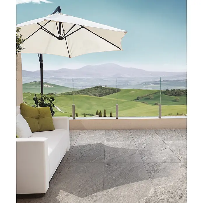 Feinsteinzeug in grauer Steinoptik 60x60, 20 mm, rutschhemmend -  - Graue gemusterte Bodenfliesen auf einer Außenterrasse mit Sofa und Sonnenschirm, mit Blick auf eine Hügellandschaft.