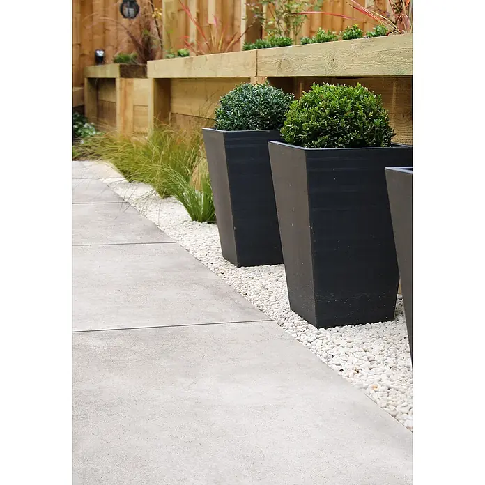 Feinsteinzeug in gewachster weißer Betonoptik 60x120 - Italienische Fliesen -  - Graue Betonplatten bilden einen modernen Gartenweg, gesäumt von weißen Ziersteinen und schwarzen Pflanzgefäßen.