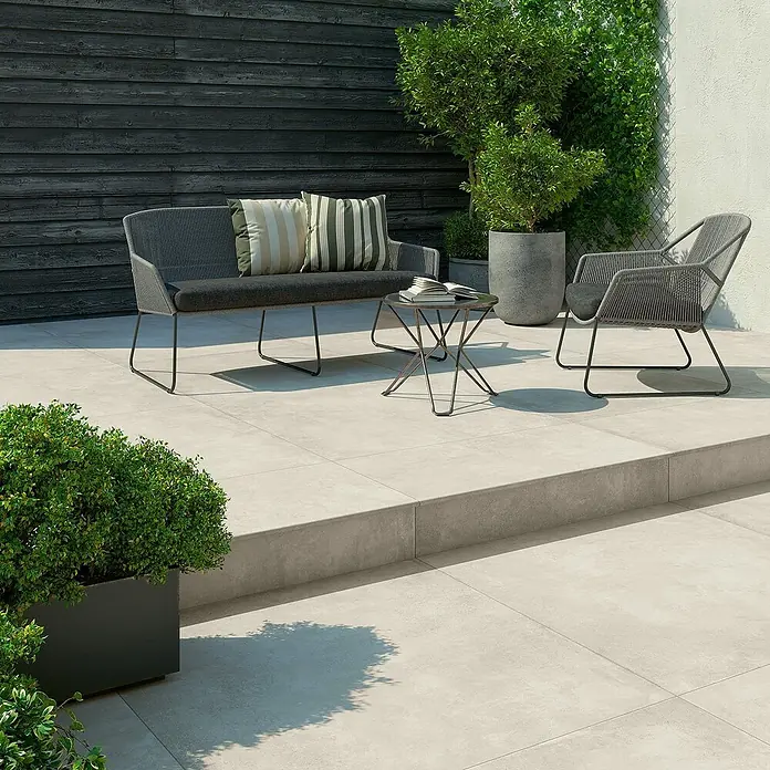 Feinsteinzeug in gewachster weißer Betonoptik 60x120 - Italienische Fliesen -  - Eine einladende Terrasse mit hellbeigen Fliesen, einer dunkelgrau geflochtenen Bank, einem Sessel mit gestreiften Kissen und einem kleinen Beistelltisch.