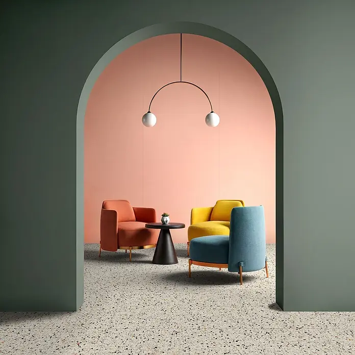 Feinsteinzeug in farbiger salbeigrüner Betonoptik 60x60 - Ital. Fliesen -  - Moderne Lounge mit bunten Sesseln, schwarzem Beistelltisch, Terrazzoboden und einer markanten rosa Wand, umrahmt von einem Torbogen.