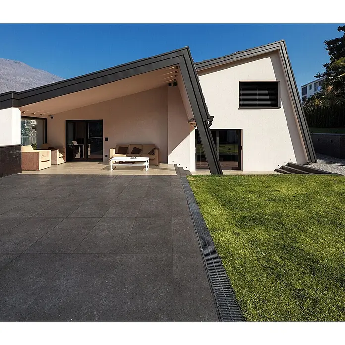 Feinsteinzeug in anthrazit Betonoptik 60x60 - Italienische Fliesen -  - Dunkelgraue, quadratische Pflasterplatten auf einer Terrasse neben einem Haus, getrennt durch eine lineare Entwässerungsrinne vom grünen Rasen.