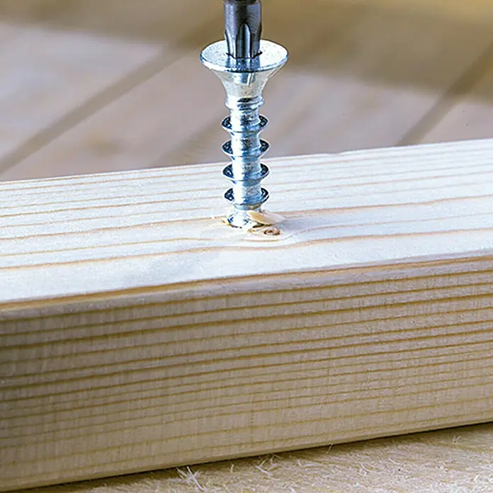 Profi Depot Tornillo tirafondo VZ - 4,5 x 50 mm, 100 ud., Zincado, TX para accionamientos TORX - Primer plano de un tornillo que se introduce en un trozo de madera con un destornillador, creando virutas de madera.