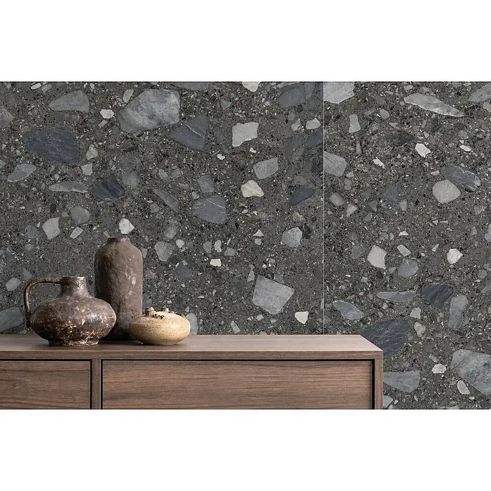 Feinsteinzeug in schwarzer Steinoptik 120x120 - Italienische Fliesen -  - Dunkelgraue Terrazzo-Wand mit hell- und dunkelgrauen Steinsplittern hinter einem Holzschrank mit drei Keramikvasen.
