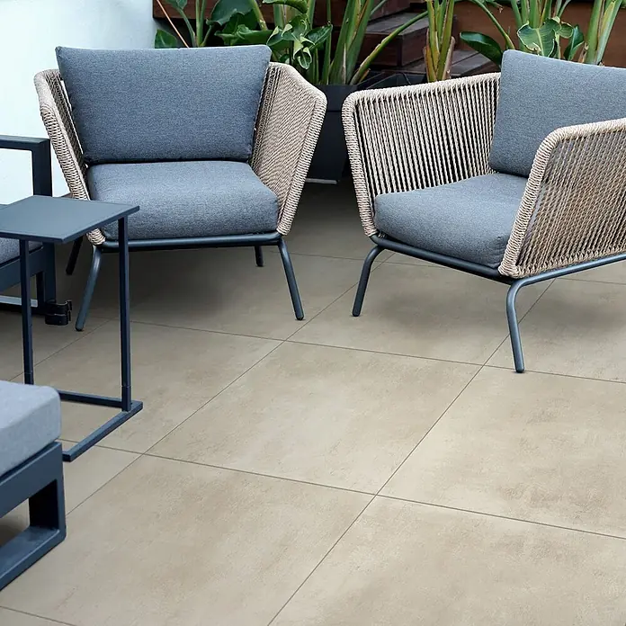 Feinsteinzeug in elfenbein Harzoptik 60x120 - Italienische Fliesen -  - Zwei beige geflochtene Outdoor-Sessel mit grauen Kissen und ein dunkelgrauer Beistelltisch auf hellbraunen Terrassenfliesen.