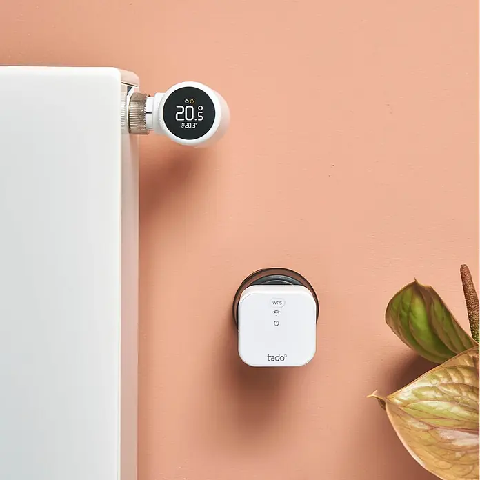 Tado Controlador inteligente Bridge X - Blanco, L x An x Al: 3,1 x 6 x 6 cm - Un termostato inteligente blanco para radiadores, que muestra «20.9°C», está fijado a un radiador, junto a un puente de internet tado blanco enchufado a una pared melocotón.