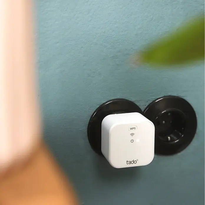 Tado Juego de termostato + Bridge X - Blanco/Negro, Termostáto + Bridge - Dispositivo tado° blanco enchufado a una toma de corriente negra en una pared azul verdosa.