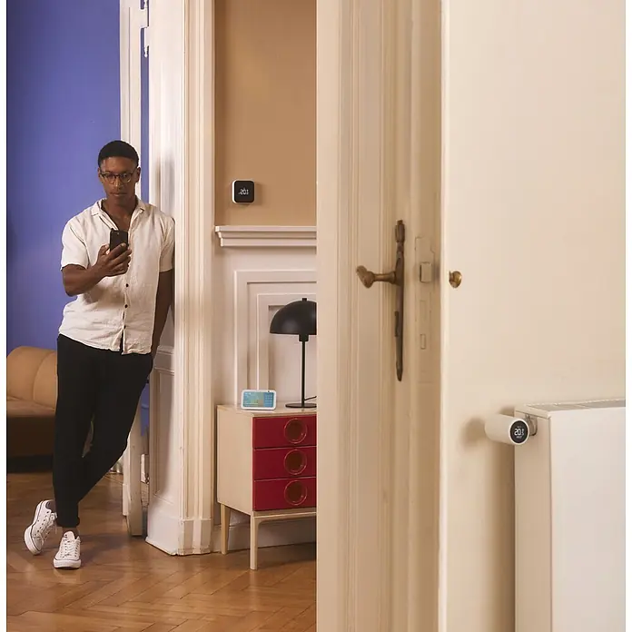 Tado Termostato de radiador Cabezal X Quattro - Blanco, 4 ud. - Hombre usa smartphone junto a dispositivos de hogar inteligente: termostato de pared, pantalla inteligente y termostato de radiador, todos mostrando 20°C.