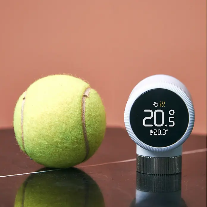 Tado Juego de termostato Bridge X + Cabezal Termoestático X - Blanco, Cabezal y Bridge - Termostato inteligente blanco con pantalla '20.5' y '20.3°' junto a una pelota de tenis para escala.