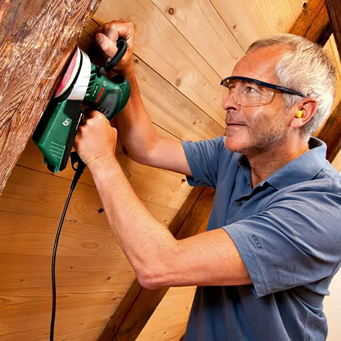 Bosch Exzenterschleifer PEX 400 AE - 350 W, Durchmesser Schleifteller: 125 mm - Ein Mann benutzt einen Bosch Mauernutfräse an einer Holzwand, ausgestattet mit Schutzbrille und Ohrstöpseln, für seine Sicherheit.