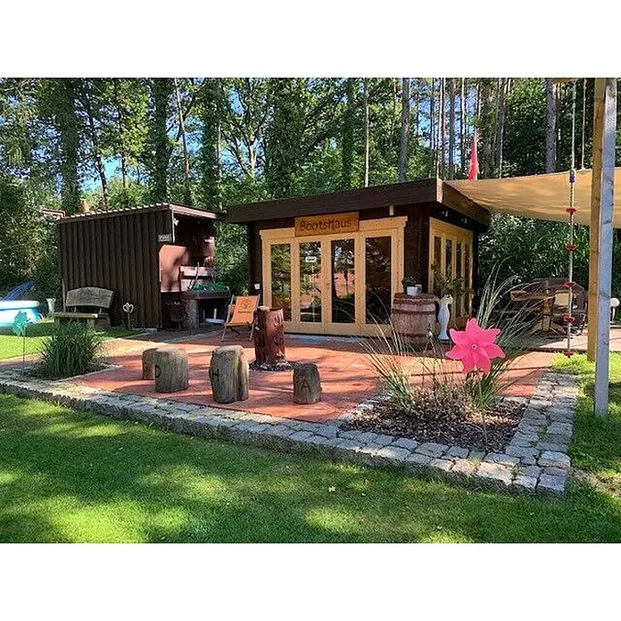 Fjordholz Gartenlounge Modell Sandra 44 A, 13.78  m², Isolierverglasung -  - Ein rustikales Holz-Bootshaus und ein angebauter Schuppen mit Terrasse in einem üppigen Garten.