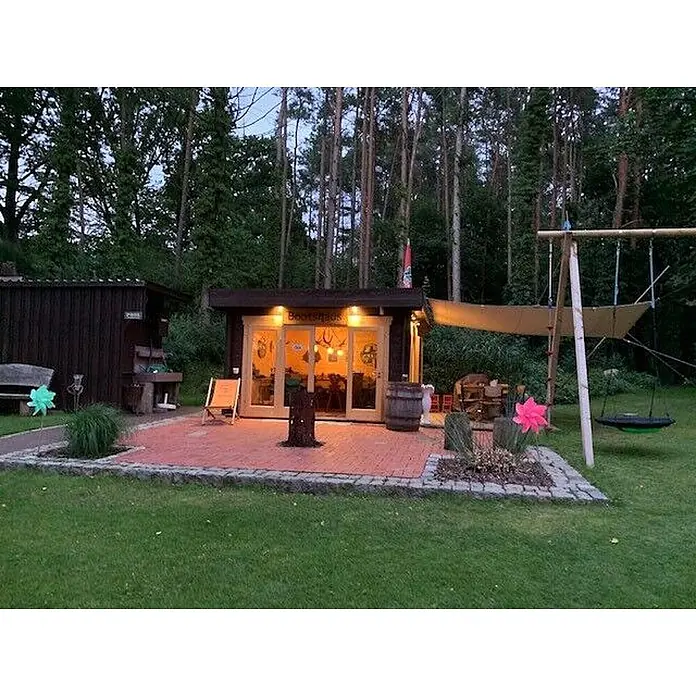 Fjordholz Gartenlounge Modell Sandra 44 A, 13.78  m², Isolierverglasung -  - Eine beleuchtete „Bootshaus“-Hütte in der Dämmerung, mit Terrasse, Sitzgelegenheiten im Freien und einer Schaukel, umgeben von Wald.