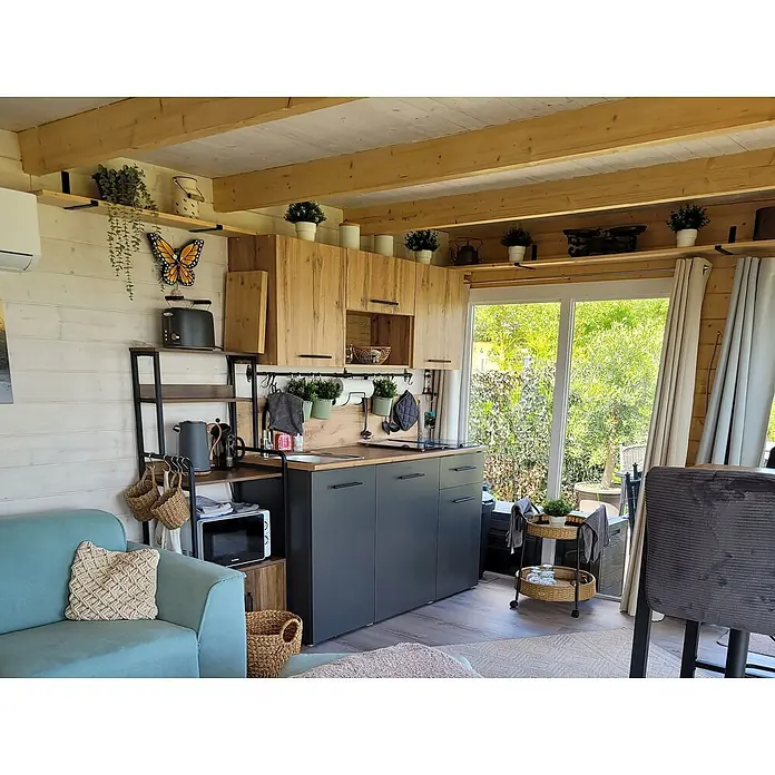 Fjordholz Gartenlounge  Lina 44 B, 29.21 m², Kiefer/Pinie -  - Kompakte dunkelgraue Küche mit hölzernen Oberschränken, Spüle, Kochfeld, integriert in ein gemütliches Wohnzimmer mit offenem Regal.