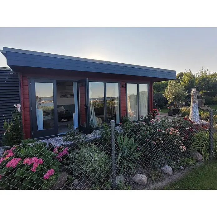 Fjordholz Gartenlounge  Lina 44 B, 29.21 m², Kiefer/Pinie -  - Modernes rotes Gartenhaus mit großen Glastüren und Fenstern, in einem blühenden Garten mit Leuchtturm.