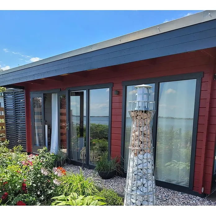 Fjordholz Gartenlounge  Lina 44 B, 29.21 m², Kiefer/Pinie -  - Dekorative Gabionen-Leuchtturm-Gartendeko mit Steinen neben einer Hütte.