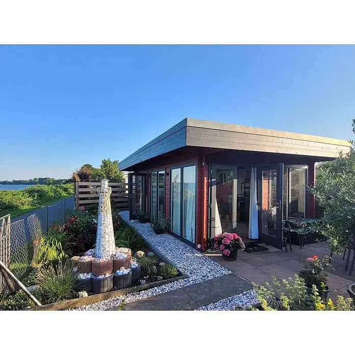 Fjordholz Gartenlounge Modell Lina 44 B, 29.21 m², Farbbehandlung Lichtgrau -  - Ein modernes rotbraunes Gartenhaus mit großen Glastüren, Flachdach, umgeben von einem Garten mit Leuchtturm und Wasserblick.