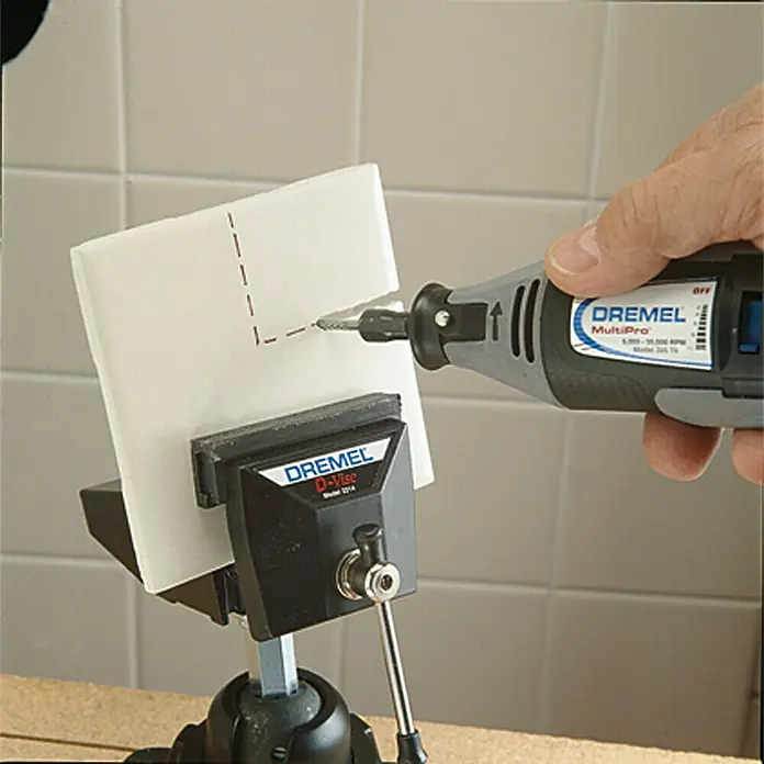 Dremel Fliesen-Fräsmesser Mod. 562 - Arbeitsdurchmesser: 3,2 mm - Dremel MultiPro-Rotationswerkzeug bohrt ein Loch in eine quadratische weiße Fliese, die in einem Dremel D-Vise-Schraubstock befestigt ist.