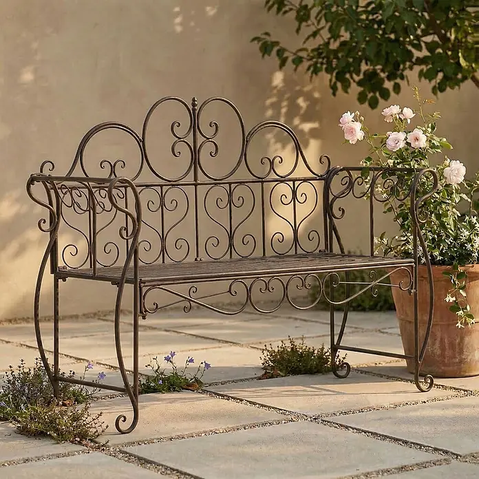 Furnicato Gartenbank Minna aus Metall -  - Eine antike dunkelbraune Gartenbank aus Schmiedeeisen mit dekorativen Schnörkeln steht auf einer Terrasse neben einem Topf mit rosa Rosen.