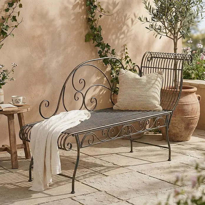 Furnicato Gartenbank Karma Jugendstil aus Eisen -  - Verzierte Gartenliege aus Schmiedeeisen mit beigem Kissen und weißer Decke auf einer Steinterrasse.