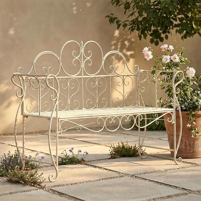Furnicato Gartenbank Minna aus Metall -  - Weiße, verzierte Gartenbank aus Schmiedeeisen mit Schnörkeln, draußen auf einer Terrasse mit Blumen platziert.