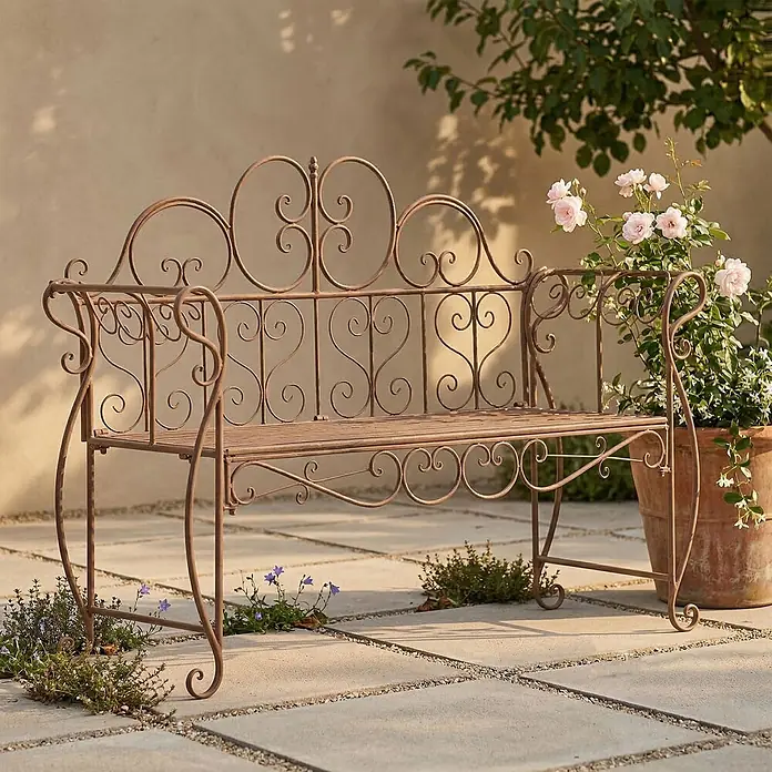Furnicato Gartenbank Minna aus Metall -  - Dekorative, rostbraune Metall-Gartenbank mit Schnörkeln auf einer Terrasse, umgeben von Rosen und Pflanzen, ideal für den Außenbereich.