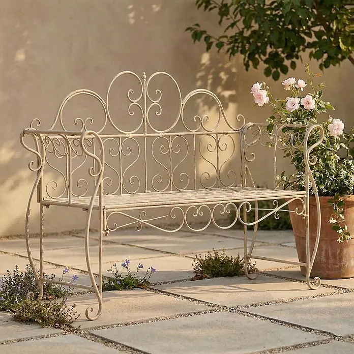 Furnicato Gartenbank Minna aus Metall -  - Verschnörkelte Gartenbank aus antikem cremefarbenem Metall auf einer Terrasse mit Blütenpflanzen.