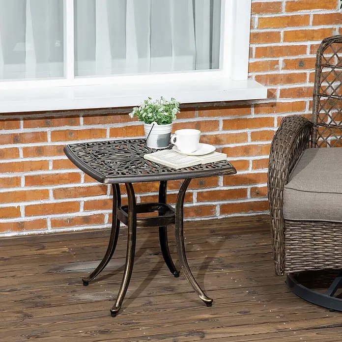 Furnicato Beistelltisch Aluminiumguss Bronze mit Schirmloch -  - Dunkler Outdoor-Beistelltisch aus Gusseisen mit verzierter Platte auf einer Holzterrasse neben einem Korbsessel, mit Pflanze, Tasse und Buch.
