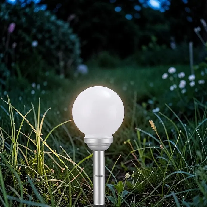 Solar Kugelleuchte Ola 15cm mit Erdspieß -  - Weiße runde Solar-Gartenleuchte mit silbernem Stab in taufeuchtem Gras bei Dämmerung.