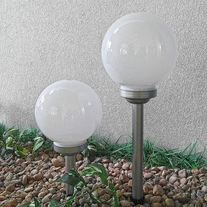Solar Kugelleuchte Ola 15cm mit Erdspieß -  - Zwei weiße kugelförmige Gartenleuchten mit silbernen Pfosten in einem Kies- und Pflanzenbeet vor einer strukturierten Wand.