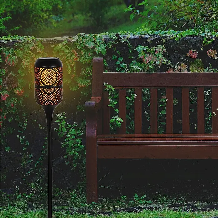 LED Solar-Wegeleuchte mit Flammeneffekt -  - Schwarze Solar-Gartenfackel mit orangefarbenem Flammeneffektlicht, beleuchtet einen Gartenweg neben einer Holzbank.