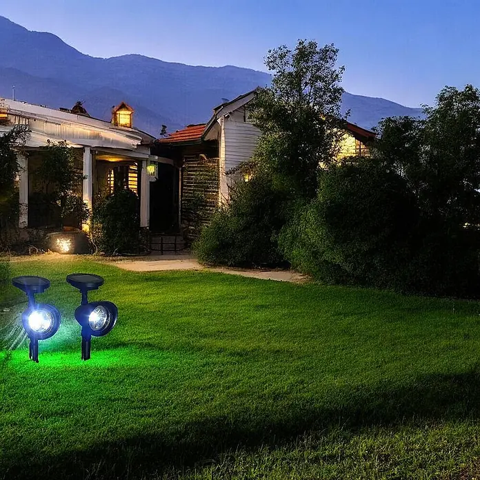2 er Set LED Solarleuchten Solarstrahler -  - Zwei dunkle Solarstrahler, einer mit weißem und einer mit grünem Licht, beleuchten einen Rasen vor einem Haus in der Abenddämmerung.