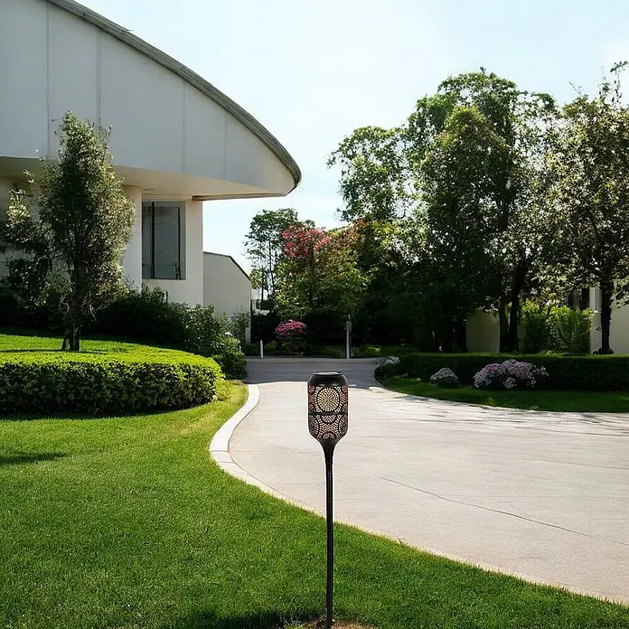LED Solar-Wegeleuchte mit Flammeneffekt -  - Schwarze Solarfackel mit ornamentalem, leuchtendem Oberteil, in einem Garten neben einer Auffahrt.