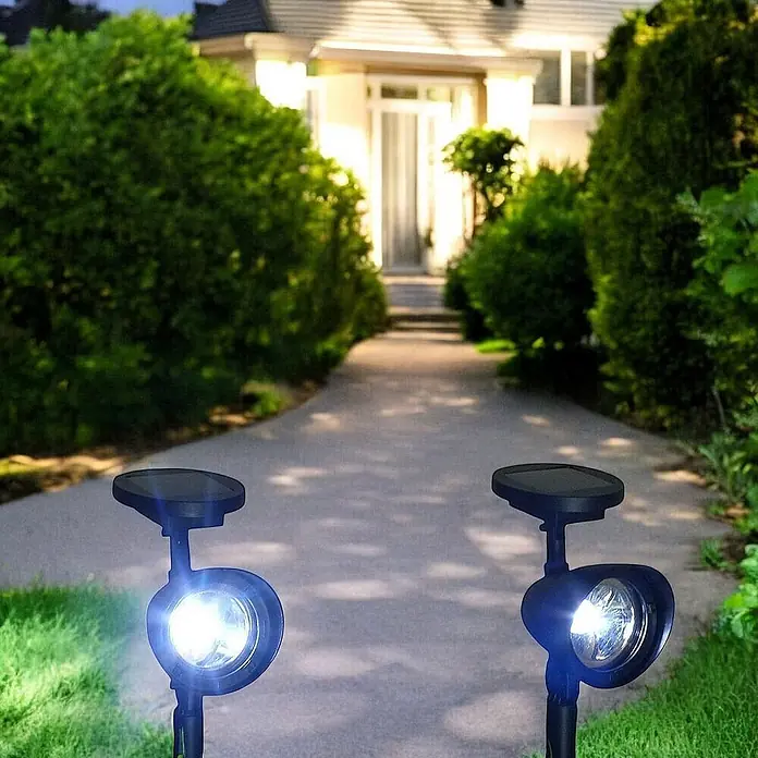 2 er Set LED Solarleuchten Solarstrahler -  - Zwei schwarze Solar-Spotlights beleuchten nachts einen Weg zu einem Haus, umgeben von grünen Hecken.