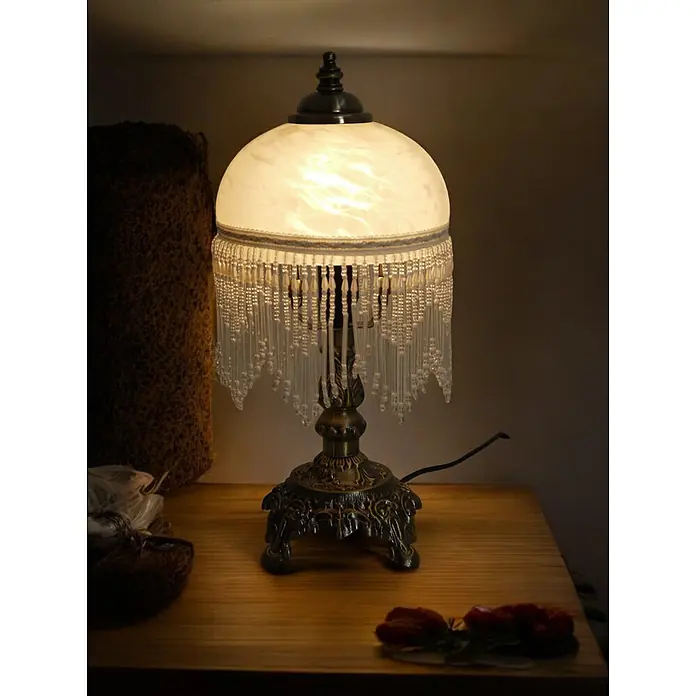 Tischleuchte Weide Landhaus Stil messing -  - Eine verzierte Tischlampe im Vintage-Stil mit beleuchtetem Milchglasschirm und Perlenfransen auf einem Holztisch.