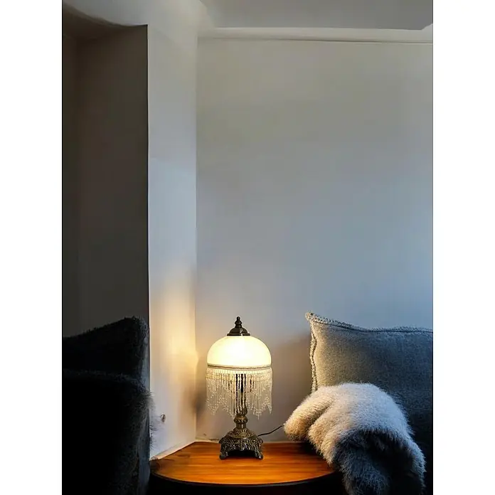 Tischleuchte Weide Landhaus Stil messing -  - Eine kunstvolle bronzene Tischlampe mit mattweißem Kuppelschirm und Perlenfransen, die warmes Licht auf einem Holztisch spendet.