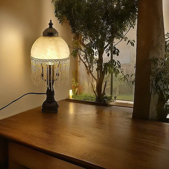 Tischleuchte Weide Landhaus Stil messing -  - Eine verzierte Tischlampe mit weißem Glasschirm und Perlenfransen, die einen warmen Schein auf einen Holztisch neben einem Fenster wirft.